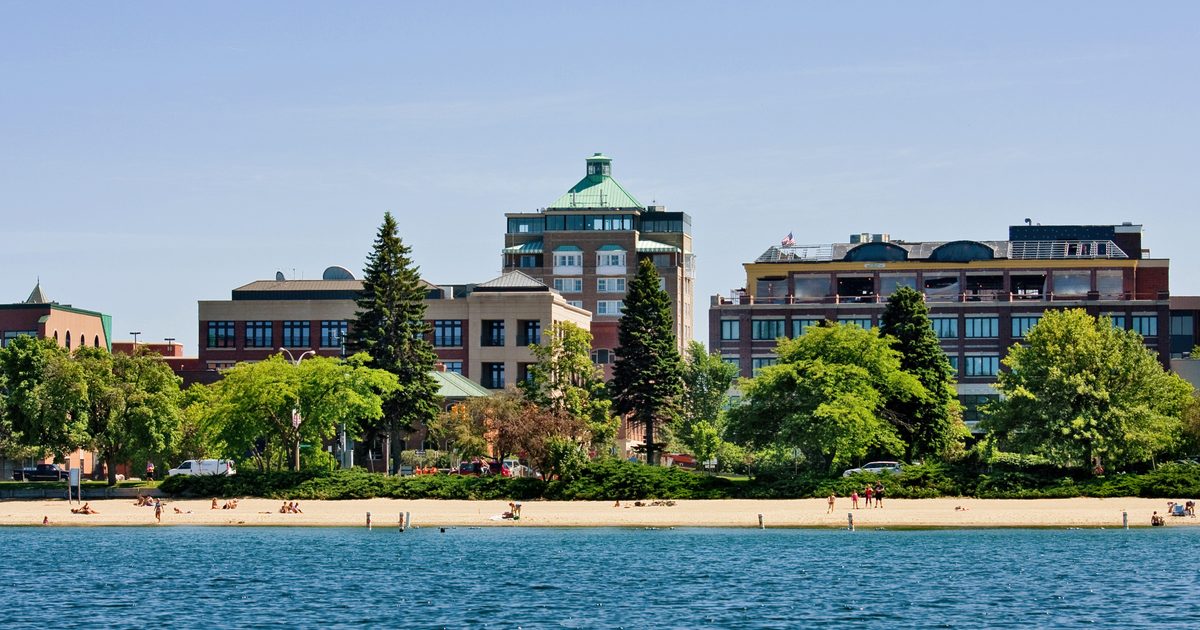 A sandy beach with people relaxing by the water, lush green trees, and several multi-story buildings in the background under a clear blue sky.