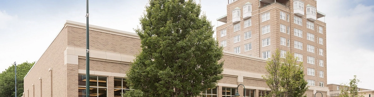 A beige brick building with large windows and a taller adjacent tower stands on a city street with parked cars, trees, and streetlights under a partly cloudy sky.