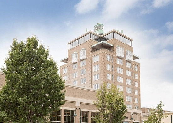 A tan brick mid-rise building with large windows, a small green-roofed structure on top, and trees in the foreground under a partly cloudy sky.