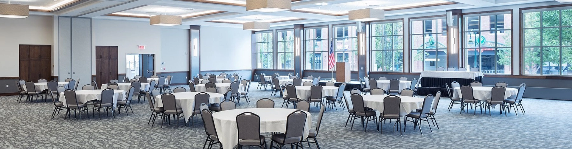 A spacious, modern banquet hall with round tables and chairs arranged throughout the room, large windows letting in natural light, and contemporary ceiling lighting.