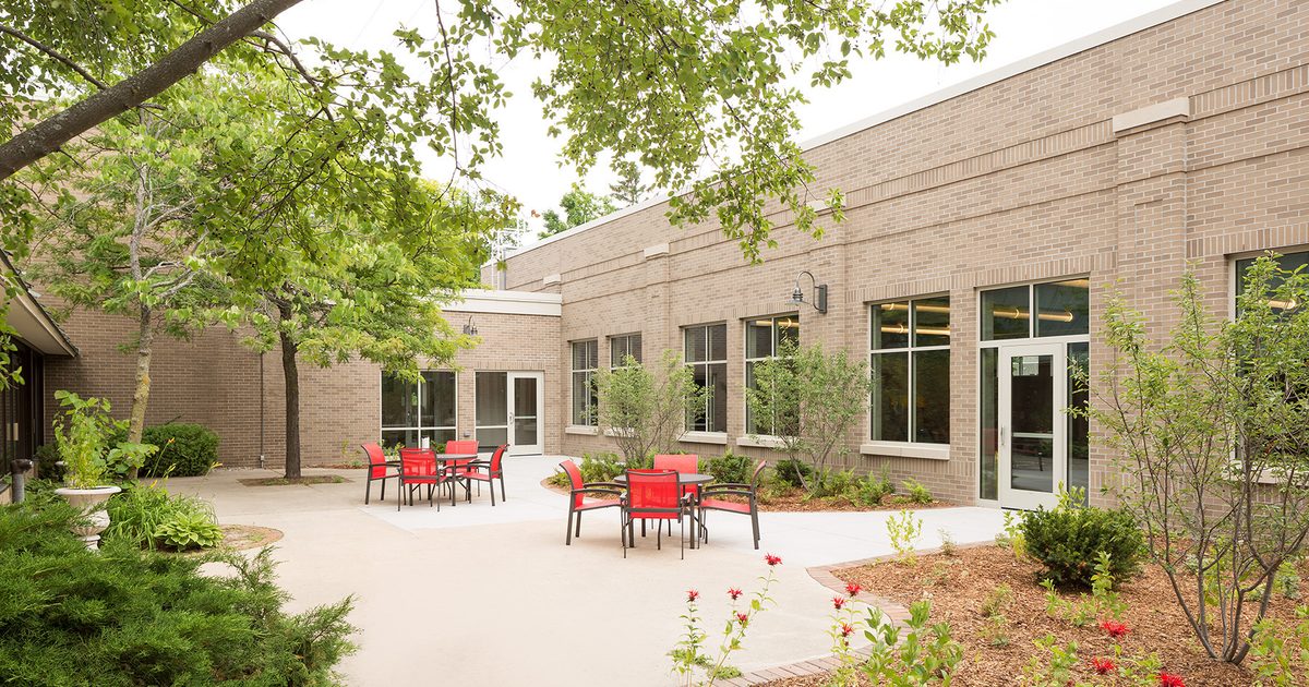 An outdoor courtyard with red chairs and tables on a concrete patio, surrounded by trees, shrubs, and a brick building with large windows and glass doors.