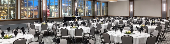 A spacious banquet hall set for an event, featuring round tables with white tablecloths, black napkins, elegant place settings, and centerpieces. Large windows line the walls, and soft lighting creates an inviting atmosphere.