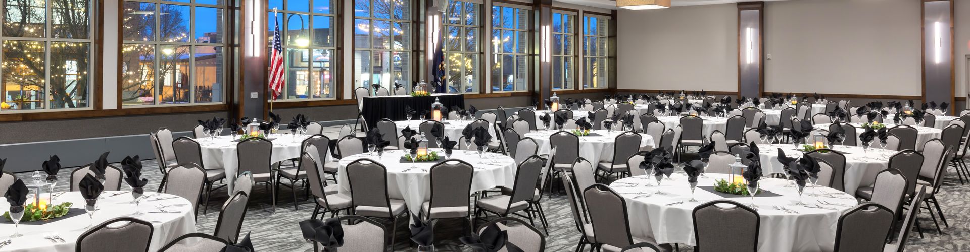 A large meeting room with round tables set for a formal event, with a view of the outside through large windows.