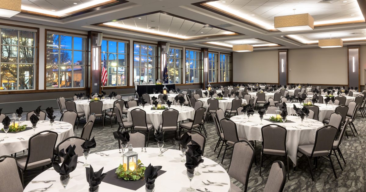 Elegant banquet hall set for an event, featuring round tables with white tablecloths, black napkins, centerpieces, and neatly arranged chairs. Large windows show evening scenery outside.