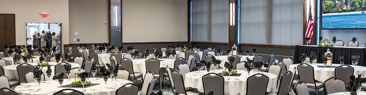 Elegant banquet hall set for an event, featuring round tables with white tablecloths, black napkins, and centerpieces. Large windows, American flag, and projection screen are visible, with people gathered near the entrance.