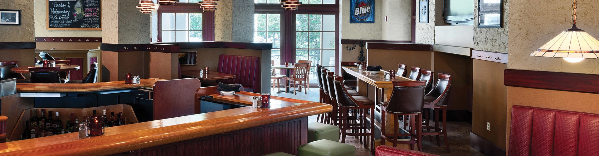 A cozy restaurant interior with a wooden bar, red booth seating, and high tables with chairs. Pendant lights hang from the ceiling, and large windows let in natural light. The atmosphere is warm and inviting.