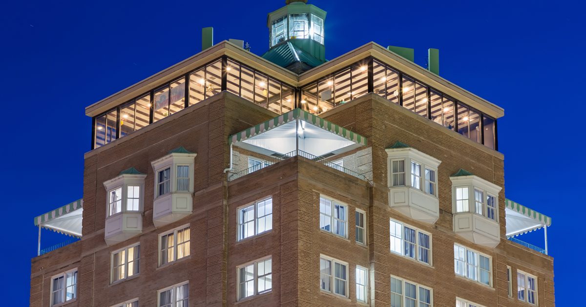A tall brick building with large windows and a glass-paneled top floor, illuminated at night against a deep blue sky. The upper section features a small lighthouse-style cupola with lights shining from within.