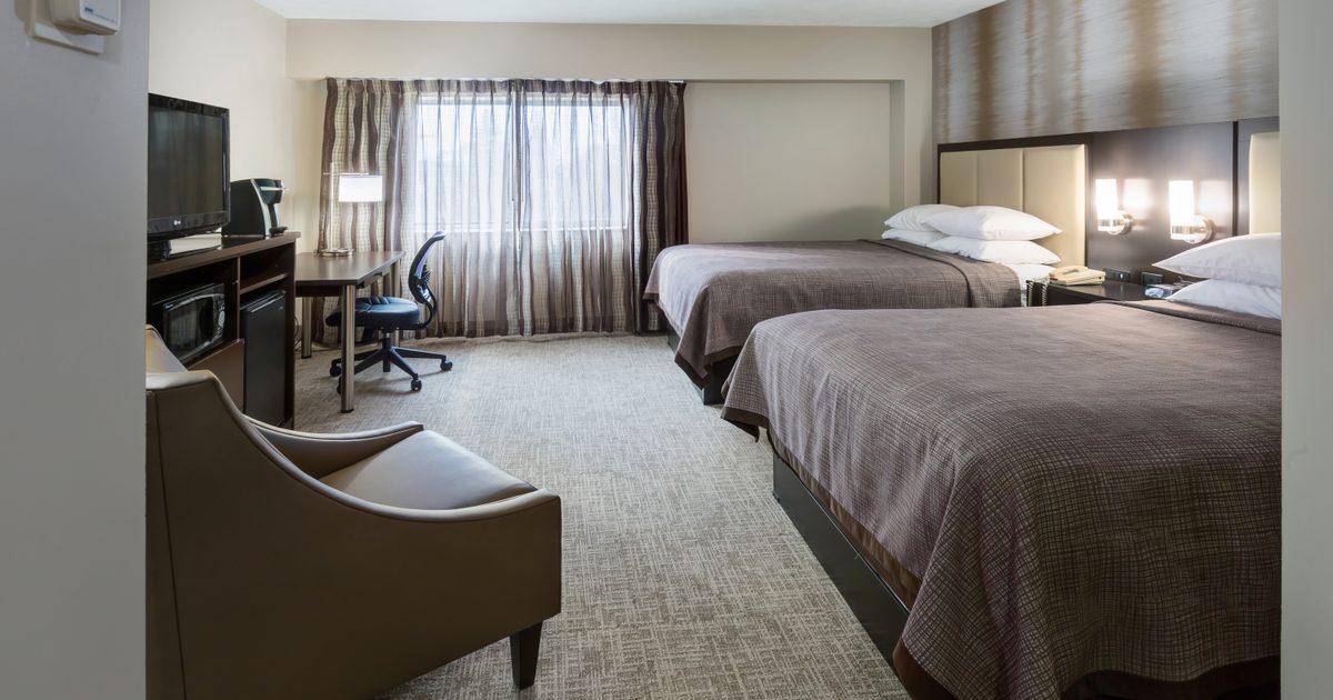 A hotel room with two double beds, a desk and chair near a window with sheer curtains, a TV on a cabinet, a microwave, a mini fridge, and an armchair on a carpeted floor.