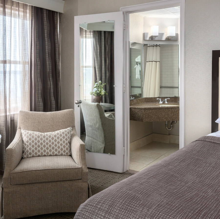 A cozy hotel room with a beige armchair, patterned pillow, round table with a floral arrangement, bed with brown cover, and an open door leading to a well-lit bathroom with a large mirror and sink.