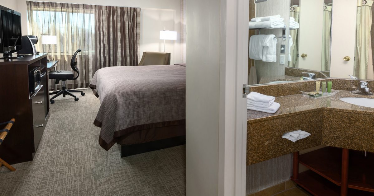 A hotel room with a bed, desk, office chair, TV, window with curtains, and an adjacent bathroom featuring a granite countertop, sink, towels, toiletries, and a large mirror.