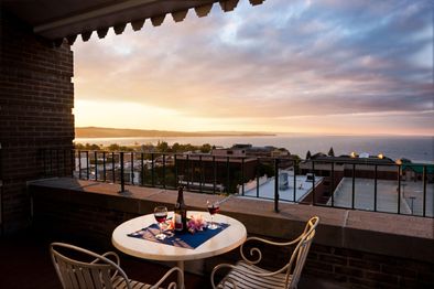 A small table with two chairs overlooks a scenic sunset view of a city and water. On the table are two glasses of wine, a wine bottle, and flowers, creating a romantic atmosphere on a balcony.
