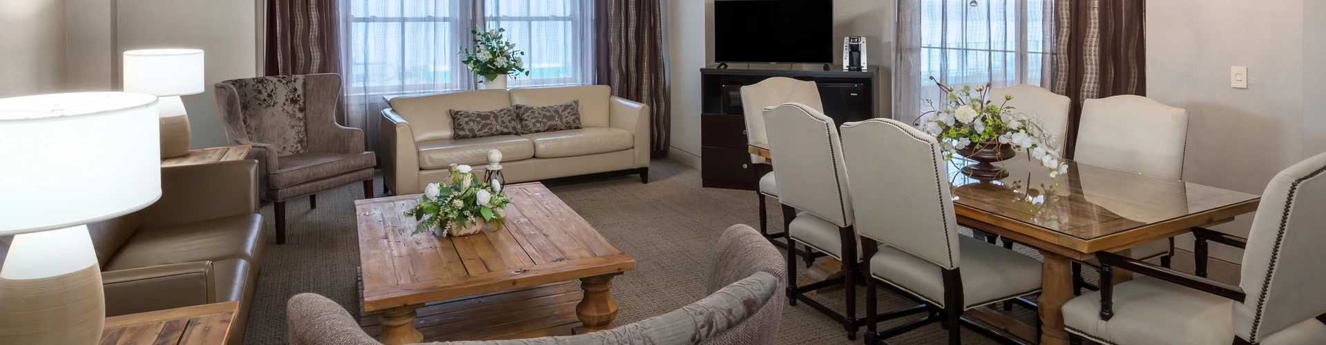 A spacious, well-lit living and dining area with beige walls, a sofa, armchairs, wooden coffee and side tables, a dining table with eight chairs, flower arrangements, a TV, and large windows with curtains.