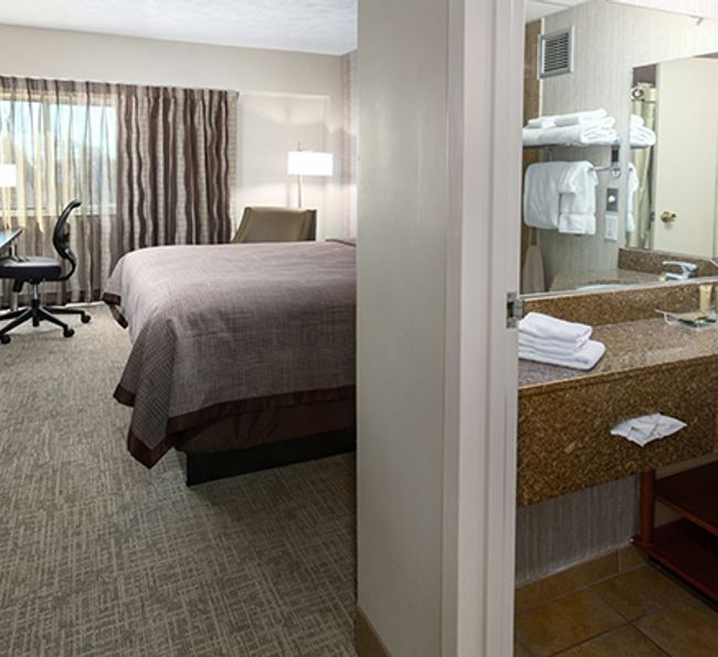 A hotel room with a bed, desk, and bathroom with a sink and mirror.