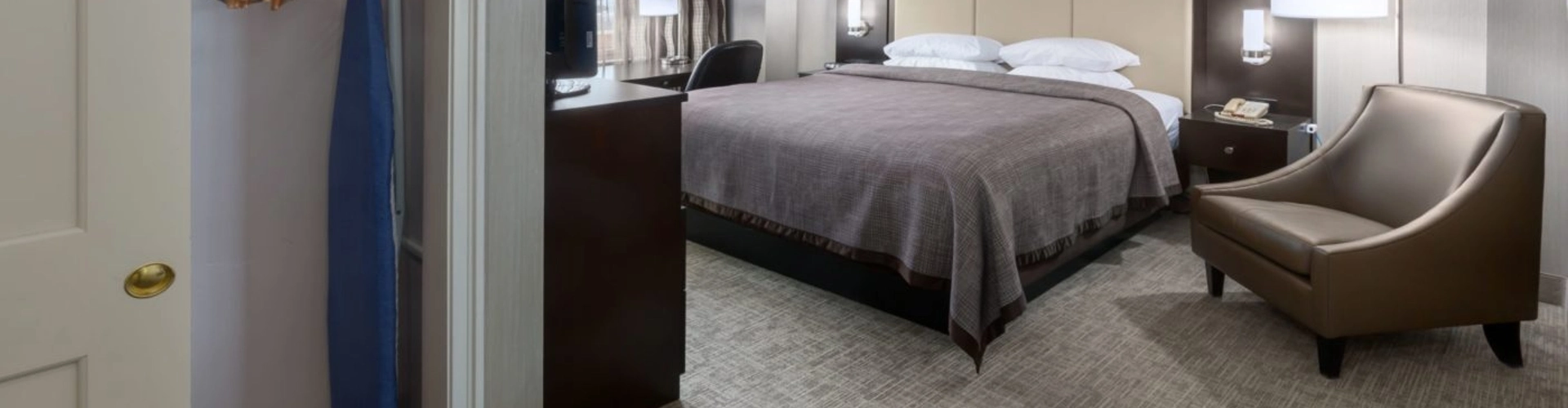 A hotel room with a neatly made king bed, brown accent chair, desk, TV, bedside lamps, and an open closet with hangers, an iron, and ironing board. Neutral tones and carpeted floor add to the cozy atmosphere.
