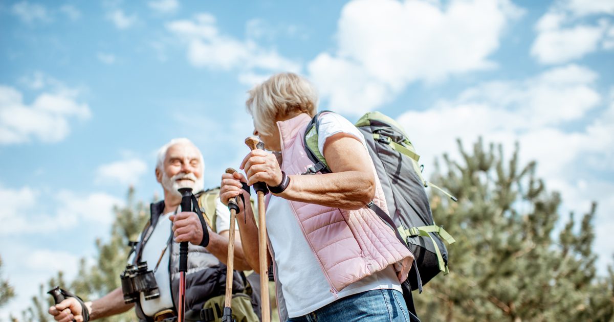 Two older adults, a man and a woman, hiking outdoors with backpacks and walking poles, smiling and enjoying a sunny day with trees and a blue sky in the background.