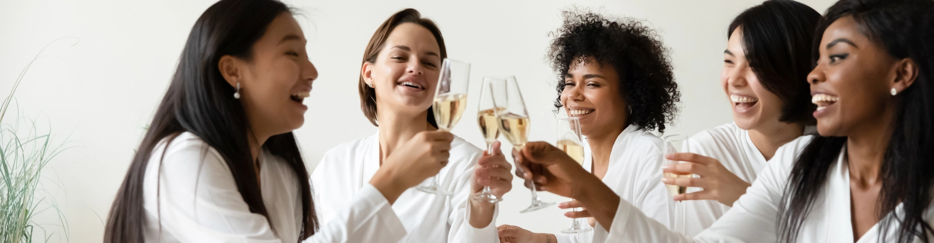 Five women in white bathrobes sit together on a bed, smiling and laughing as they toast with champagne glasses in a bright, cozy room.