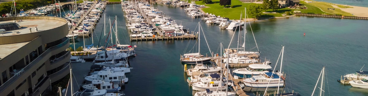 Aerial view of a marina filled with sailboats and yachts docked at piers, with clear blue-green water, nearby buildings, green trees, and a sandy beach along the shoreline under a partly cloudy sky.